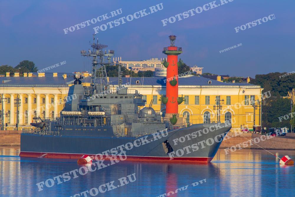 На фотографии изображен военный корабль, пришвартованный у набережной в Санкт-Петербурге. Вдалеке виднеется знаменитая Ростральная колона на Васильевском острове. Этот снимок может быть использован в материалах, посвященных Военно-морскому флоту России, городской жизни Санкт-Петербурга или как элемент декоративного оформления.